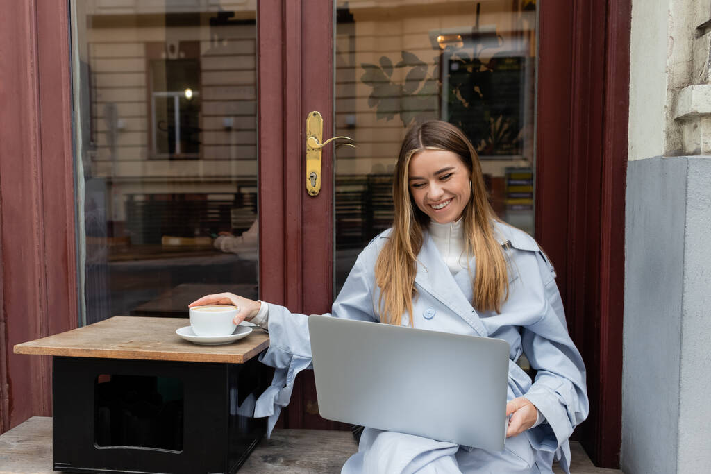 szczęśliwa kobieta w niebieskim płaszczu Trench za pomocą laptopa podczas osiągnięcia filiżanki kawy w kawiarni na świeżym powietrzu w Wiedniu   - Zdjęcie, obraz