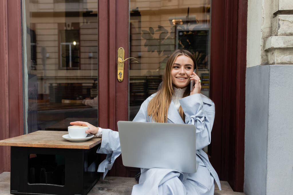 szczęśliwa kobieta w niebieskim płaszczu rozmawia na smartfonie i siedzi obok filiżanki kawy w kawiarni na świeżym powietrzu w Wiedniu  - Zdjęcie, obraz