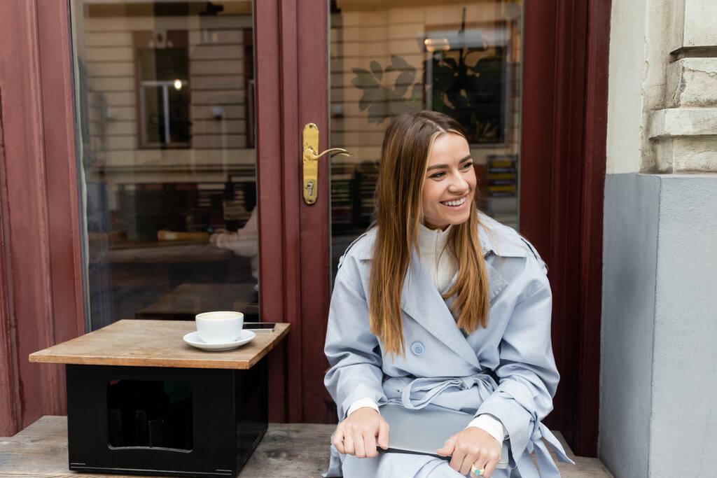 szczęśliwa młoda kobieta w niebieskim płaszczu trzyma laptopa siedząc obok filiżanki kawy w kawiarni na świeżym powietrzu w Wiedniu   - Zdjęcie, obraz