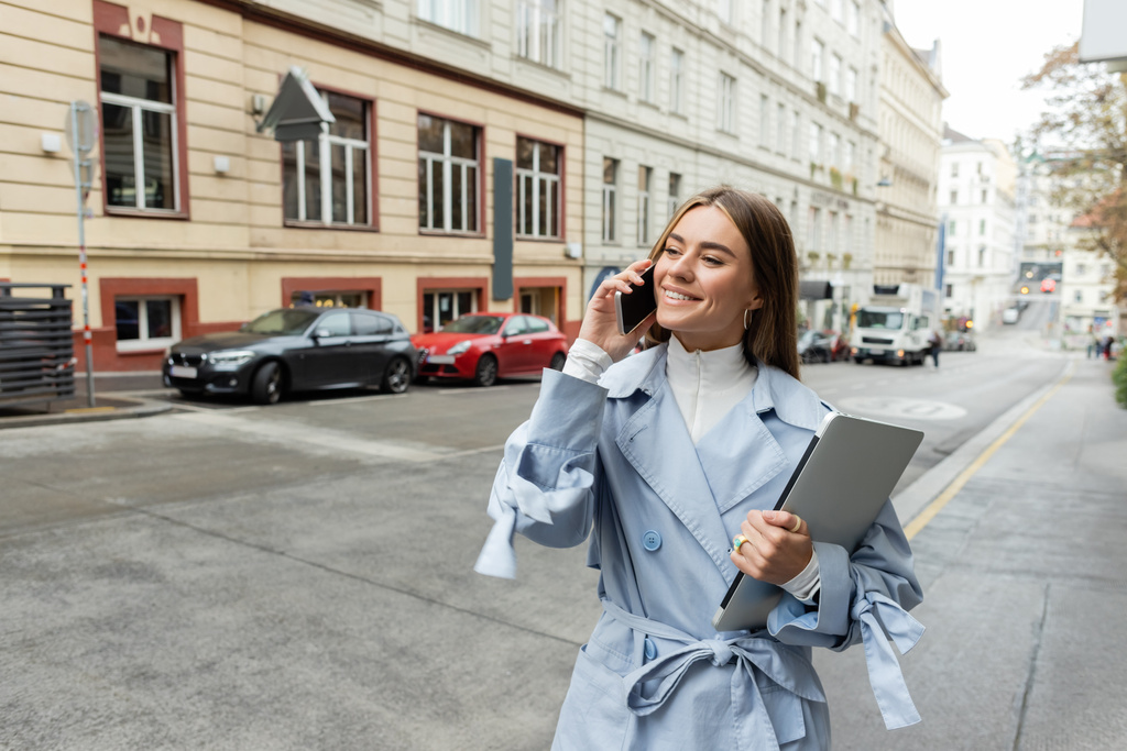 mujer alegre en gabardina azul hablando en el teléfono inteligente mientras sostiene el ordenador portátil en la calle en Viena  - Foto, Imagen