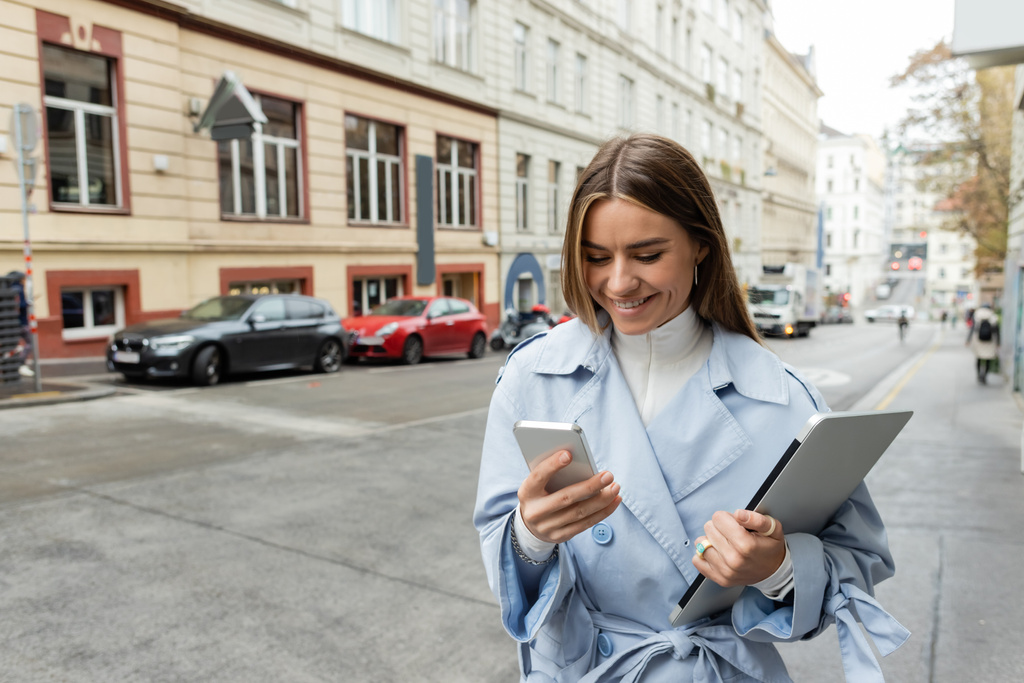 mujer alegre en gabardina azul usando teléfono inteligente mientras sostiene el ordenador portátil en la calle en Viena  - Foto, imagen