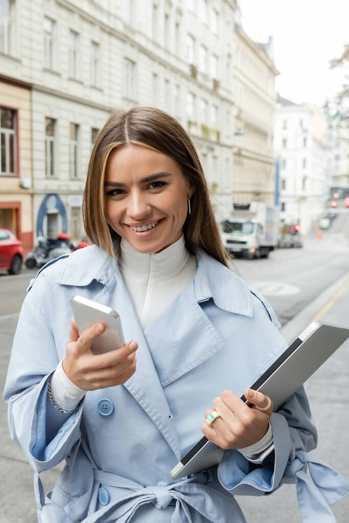 donna sorridente in trench blu con smartphone mentre tiene il computer portatile in strada a Vienna  - Foto, immagini