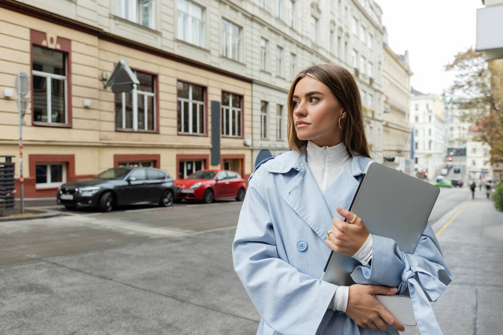 hezká žena v modrém plášti drží smartphone a notebook při chůzi na ulici ve Vídni  - Fotografie, Obrázek