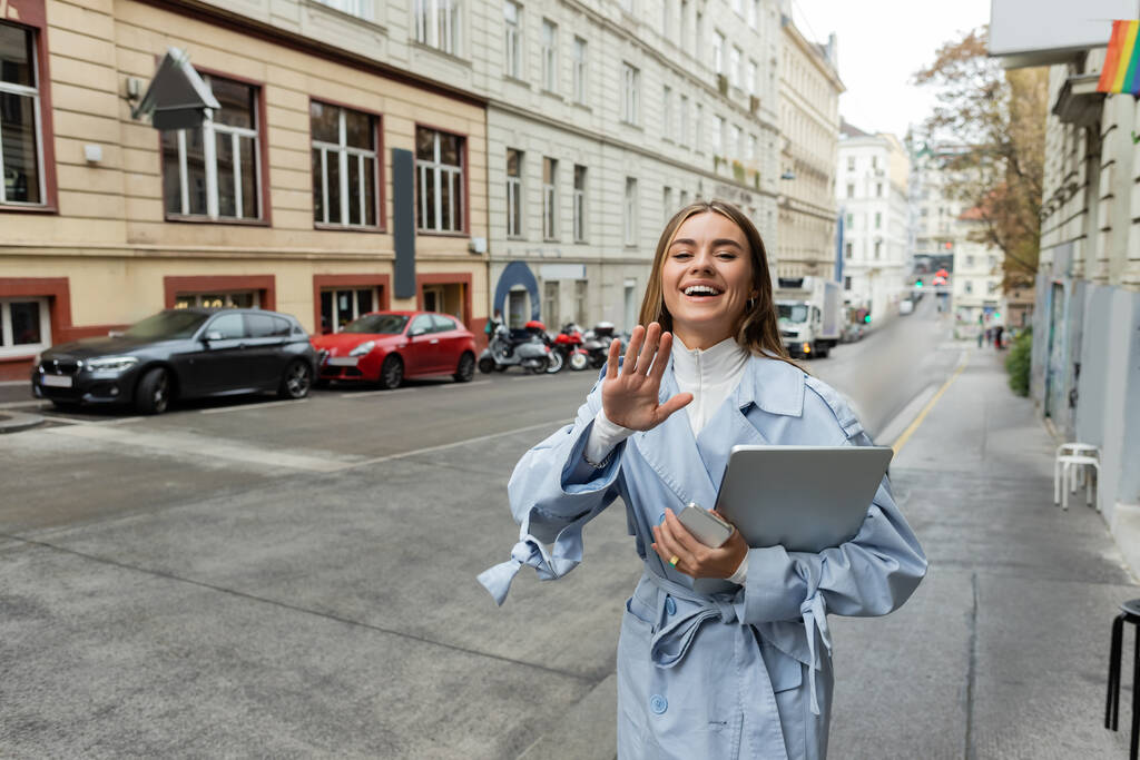 счастливая женщина в синем плаще держит смартфон и ноутбук, размахивая рукой на улице в Вене  - Фото, изображение