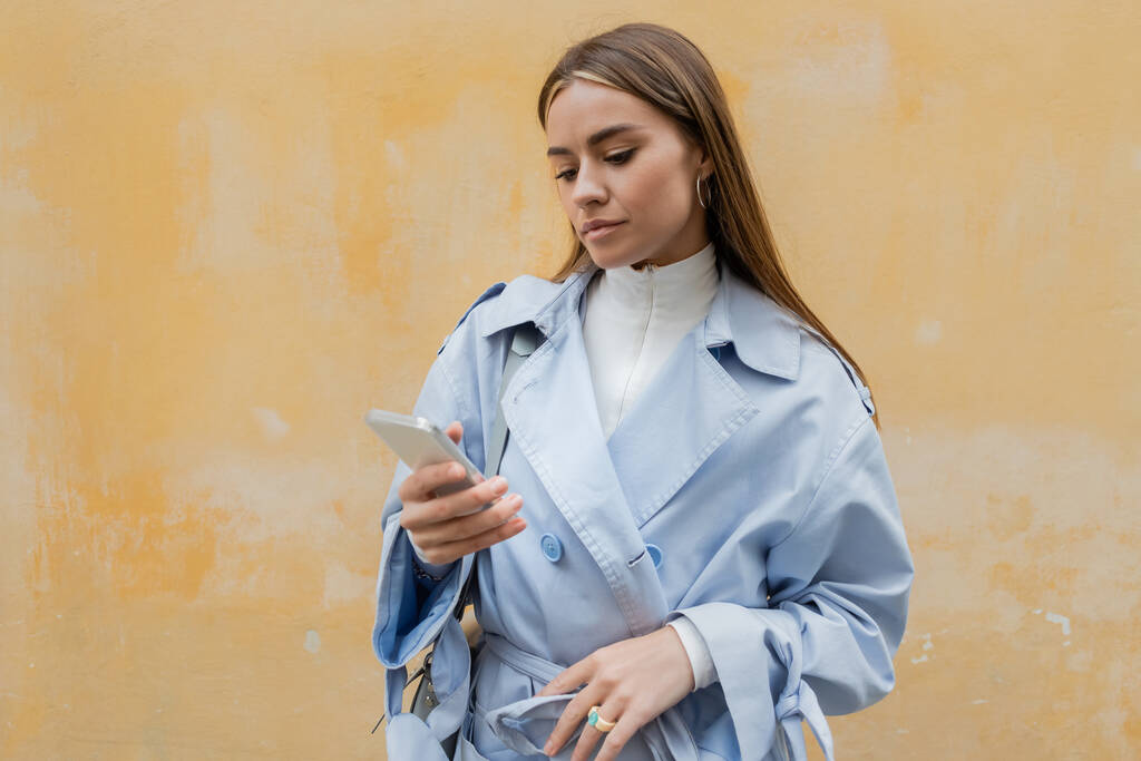 молодая женщина в синем пальто с помощью смартфона возле желтой стены на улице в Вене  - Фото, изображение
