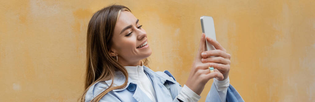 Mavi trençkotlu mutlu kadın Viyana 'da sarı duvarın yanında akıllı telefondan fotoğraf çekiyor.  - Fotoğraf, Görsel