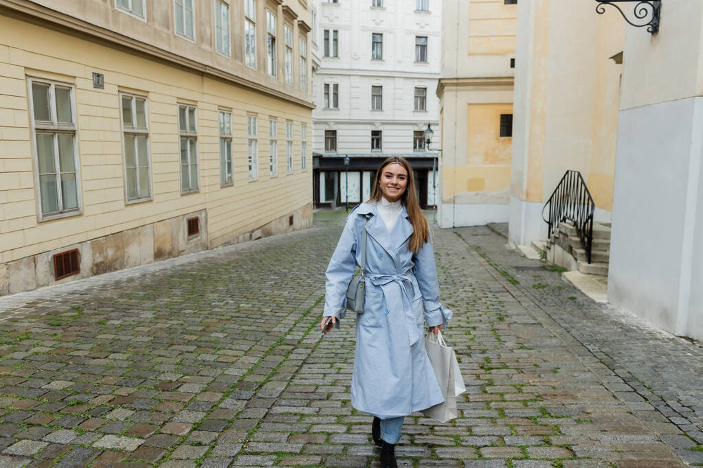 счастливая молодая женщина в синем пальто прогулка с сумками на улице в Вене  - Фото, изображение