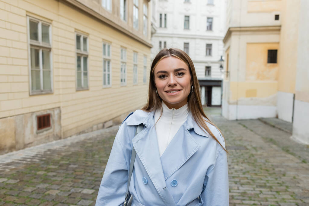 青いトレンチコートを着た陽気な若い女性がウィーンの通りの建物の近くに立っていて  - 写真・画像