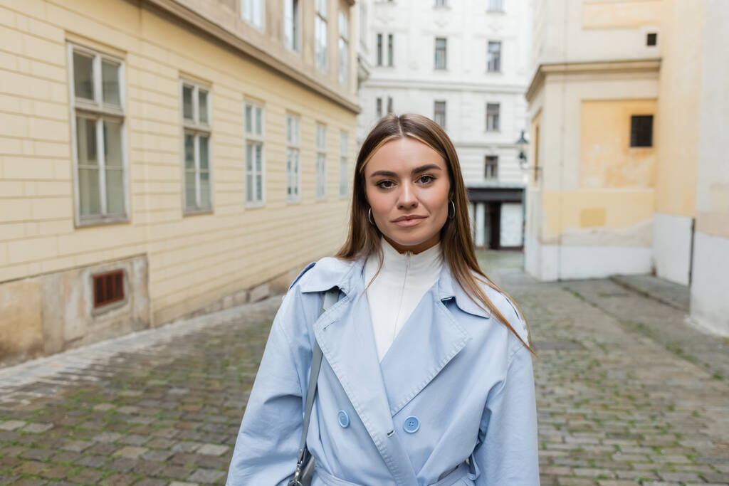 charmante jeune femme en trench-coat bleu debout près des bâtiments de la rue à Vienne  - Photo, image