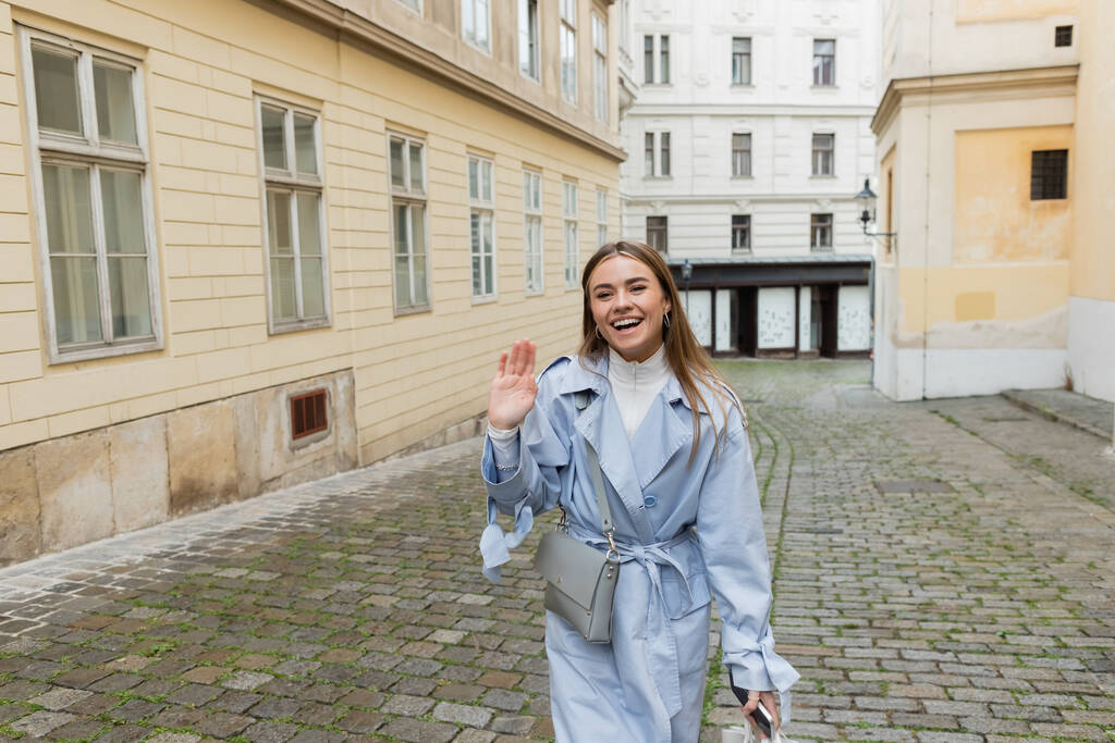 encantadora mujer en gabardina azul saludando de la mano mientras camina con un teléfono inteligente cerca de edificios en Viena  - Foto, imagen