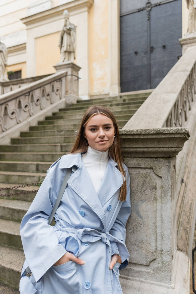 charming woman in blue trench coat standing with hands in pockets near ancient stairs on street in in Vienna  - Photo, Image
