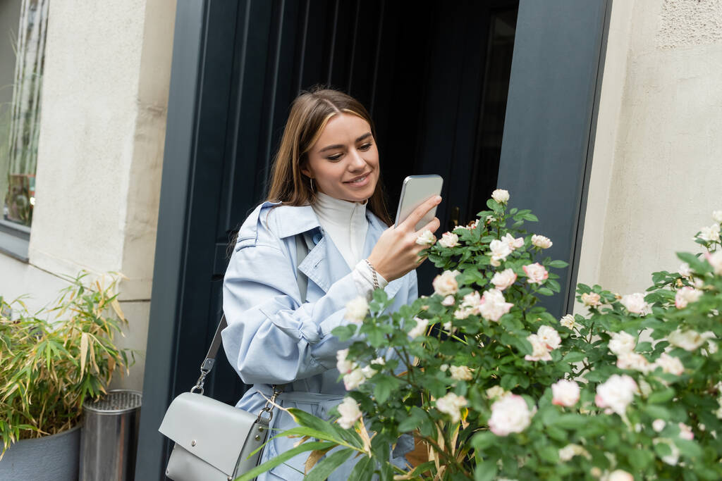 青いトレンチコートの幸せな若い女性がウィーンの通りに咲く花と緑の茂みの写真を撮る  - 写真・画像