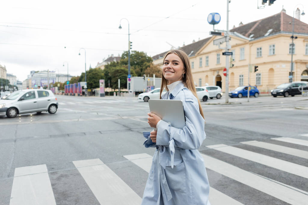 Glückliche Frau im blauen Trenchcoat hält Laptop in der Hand und überquert Straße in Wien  - Foto, Bild