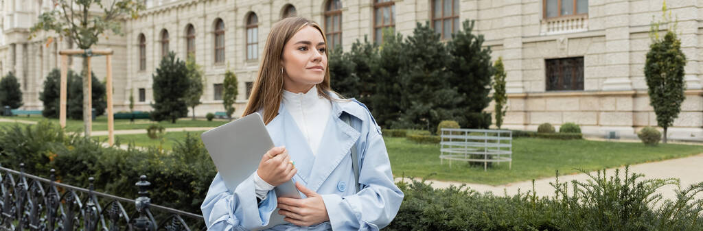 young woman in blue trench coat holding laptop near historical building in Vienna, banner  - Photo, Image