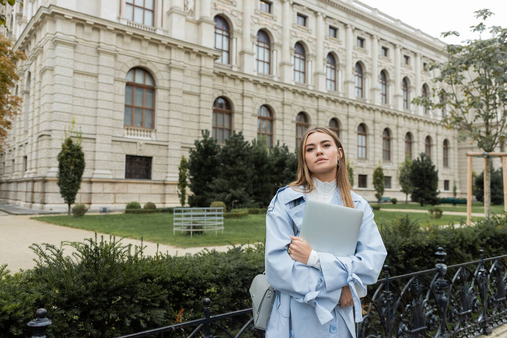 Hübsche Frau im blauen Trenchcoat mit Laptop in der Nähe historischer Gebäude in Wien  - Foto, Bild