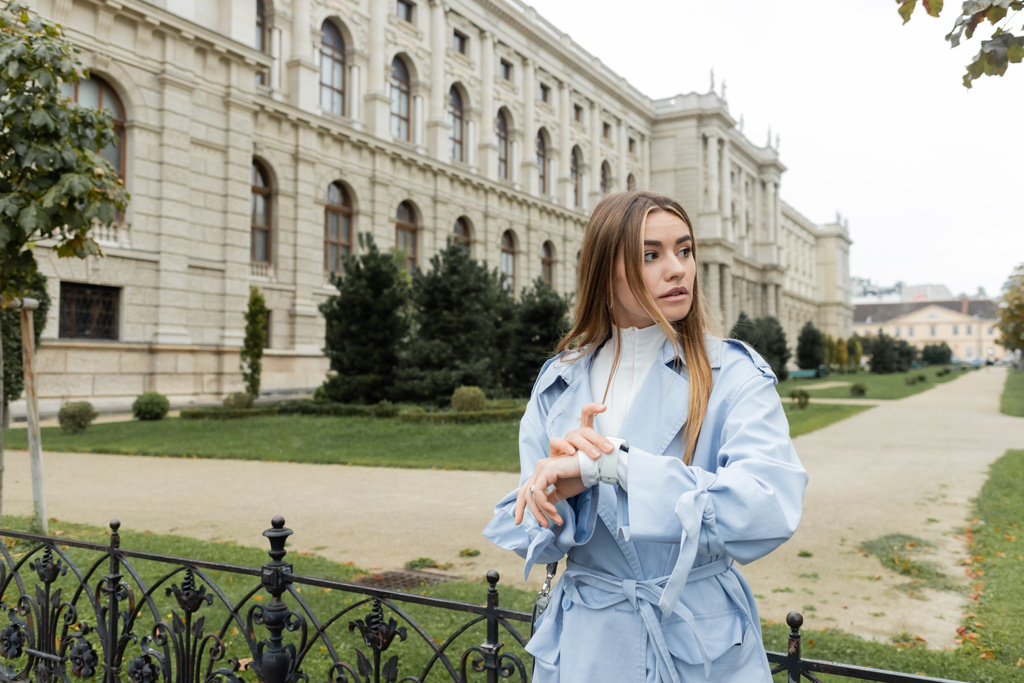 jolie femme en trench coat bleu vérifiant le temps sur la montre intelligente tout en attendant près du bâtiment historique à Vienne  - Photo, image