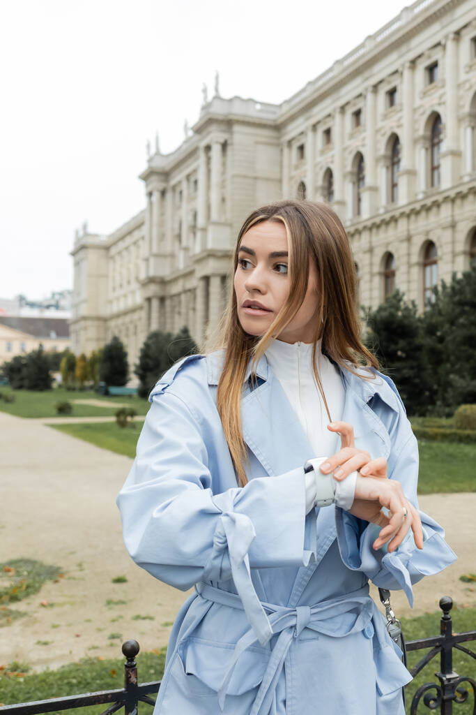 青いトレンチコートを着た若い女性がウィーンの歴史的建造物の近くで待っている間に腕時計で時間をチェック  - 写真・画像