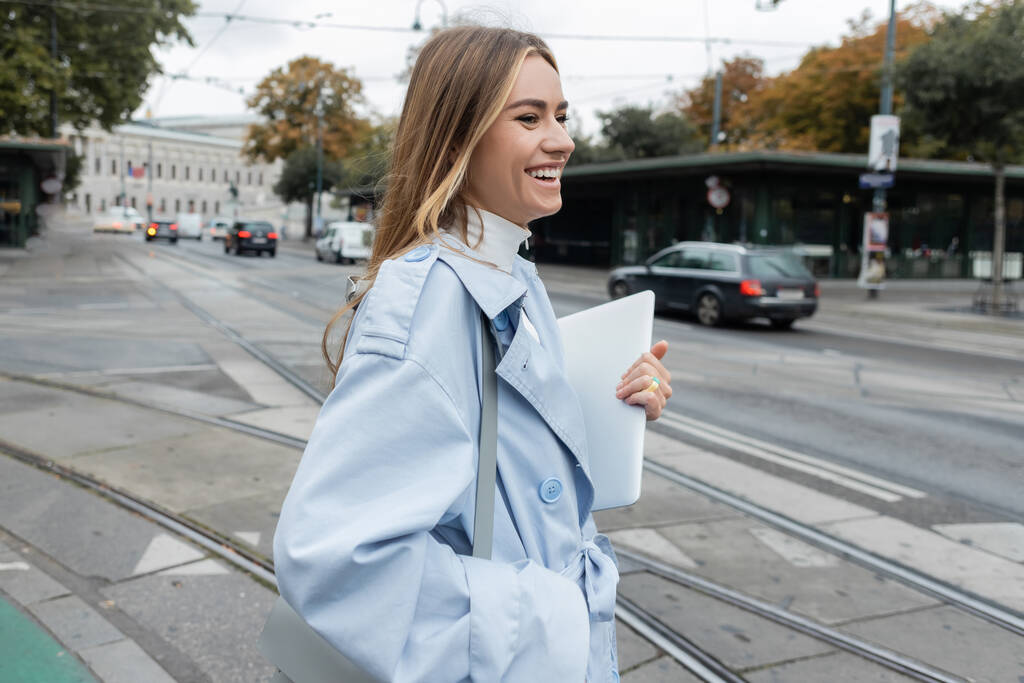 vrolijke vrouw in blauwe trench jas houden laptop tijdens het oversteken van de weg in Wenen  - Foto, afbeelding