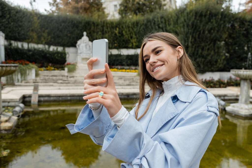 sorrindo jovem mulher em casaco de trincheira azul tirar foto no smartphone perto da fonte no parque  - Foto, Imagem