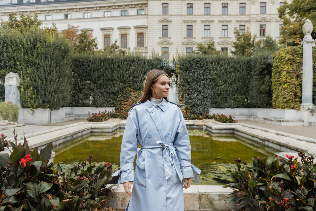jeune femme en imperméable bleu debout près de la fontaine et bâtiment historique dans le parc vert de Vienne  - Photo, image