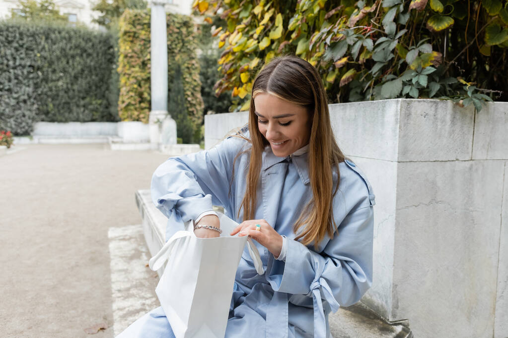 femme heureuse en trench coat bleu assis sur un banc en béton et regardant à l'intérieur du sac à provisions dans le parc vert de Vienne  - Photo, image