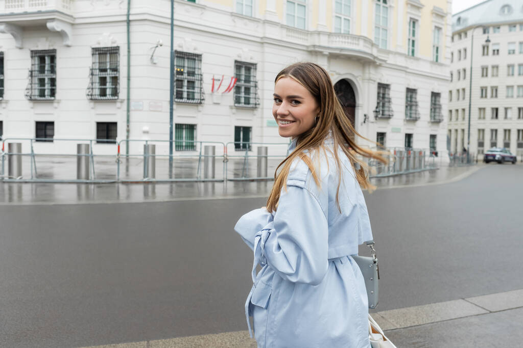 Glückliche junge Frau im blauen Trenchcoat lächelt auf der Stadtstraße in Wien  - Foto, Bild