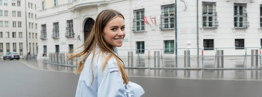 mujer joven positiva en gabardina azul sonriendo en la calle urbana de Viena, pancarta  - Foto, Imagen