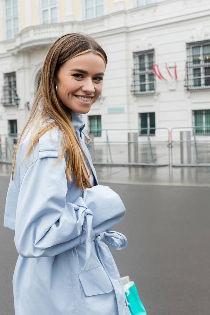 Радостная молодая женщина в голубом плаще улыбается на городской улице Вены  - Фото, изображение
