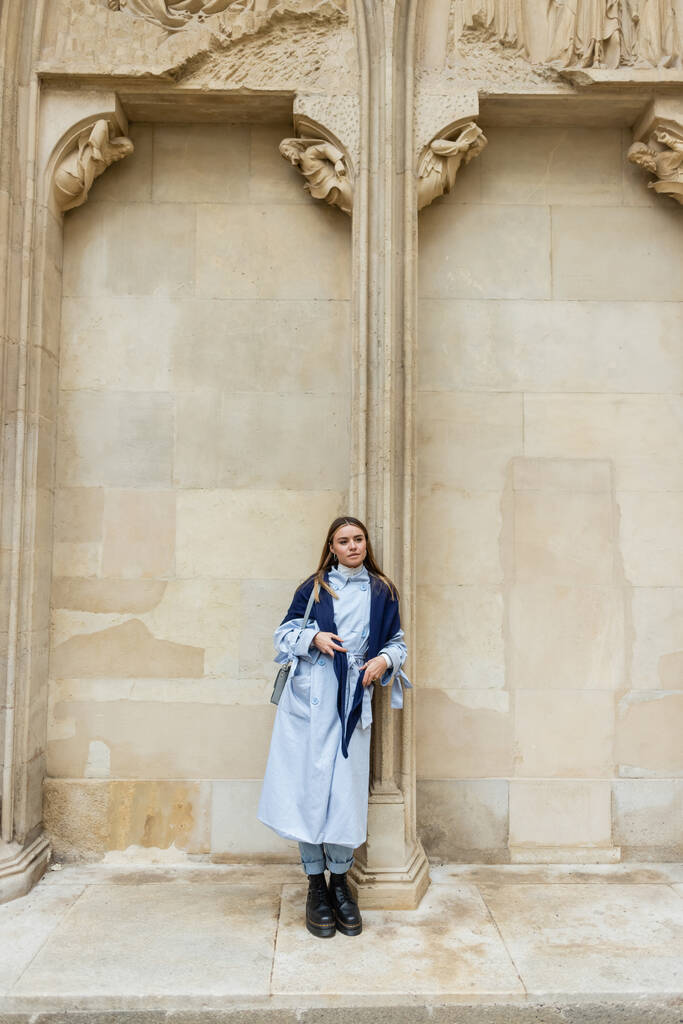 young woman with scarf on top of blue trench coat standing near historical building in Vienna  - Photo, Image