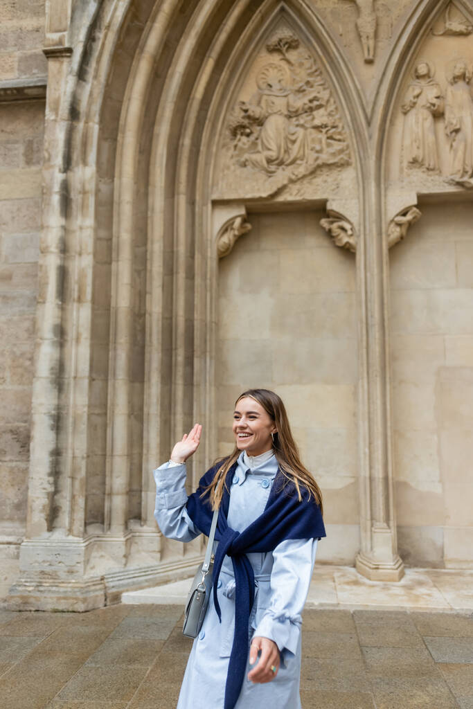 iloinen nuori nainen huivi päälle sininen trench takki heiluttaa käsi lähellä historiallista rakennusta Wienissä  - Valokuva, kuva