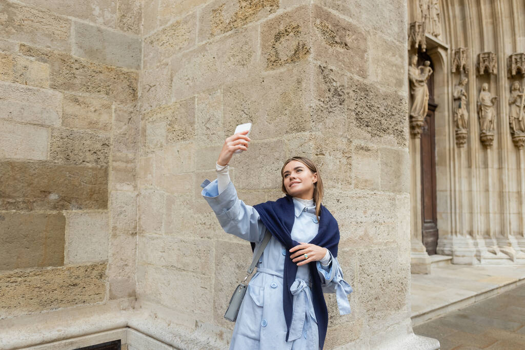 young woman with scarf on top of blue trench coat taking selfie near historical building in Vienna  - Photo, Image