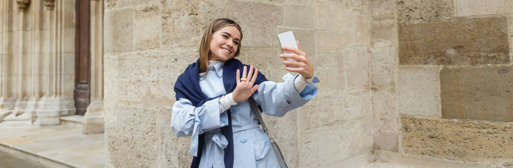 青いトレンチコートの上にスカーフを持つ幸せな女性は、ウィーンの歴史的な建物の近くにビデオ通話を持っています,バナー  - 写真・画像
