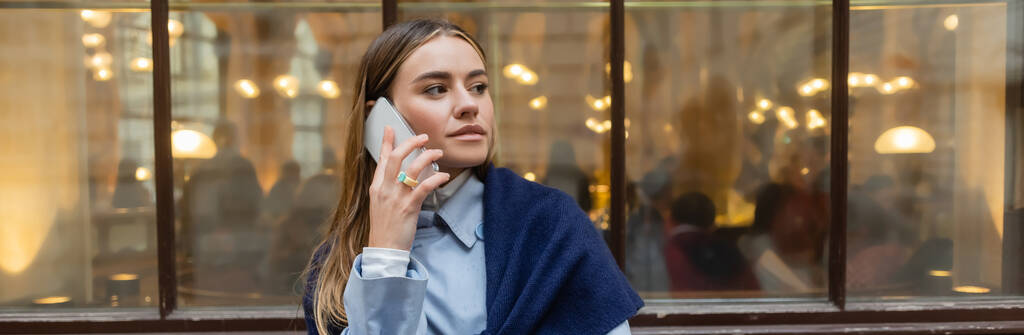 青のトレンチコートの上にスカーフを持つスタイリッシュな女性は、ウィーンの路上でスマートフォンで話して、バナー  - 写真・画像