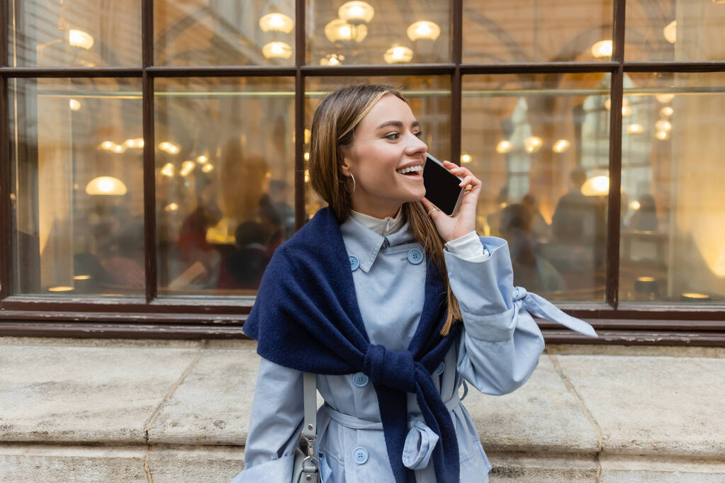 ウィーンの路上でスマートフォンで話している青いトレンチコートのスカーフを持つ陽気な女性 - 写真・画像