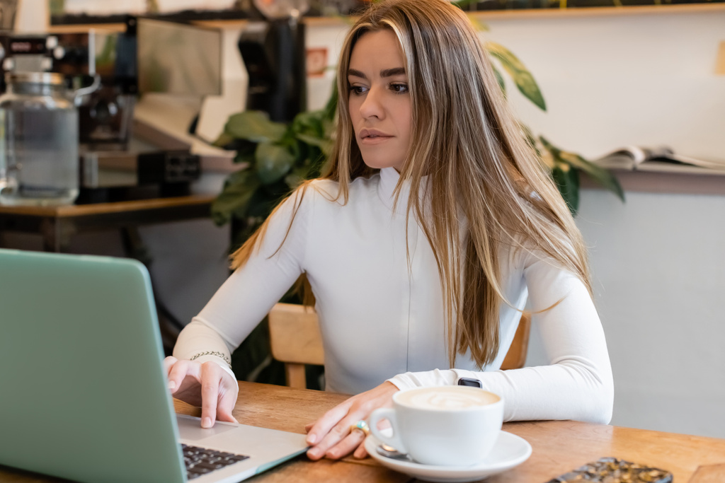 mulher encantadora em gola alta usando laptop perto de xícara de cappuccino no café  - Foto, Imagem