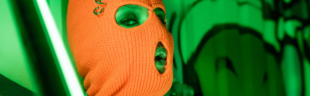 портрет сексуальной женщины в трикотажной оранжевой балаклаве возле яркой неоновой лампы и зеленой стены с граффити, баннер - Фото, изображение