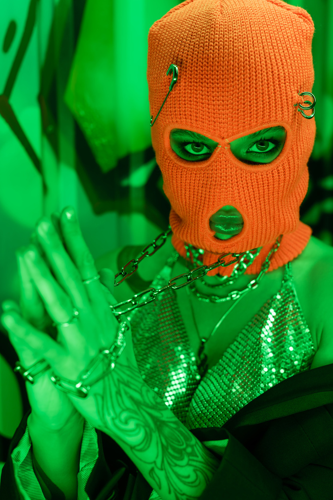 провокационная женщина в оранжевой балаклаве и металлическом топе с цепями на шее, смотрящая на камеру возле стены с граффити в зеленом свете - Фото, изображение