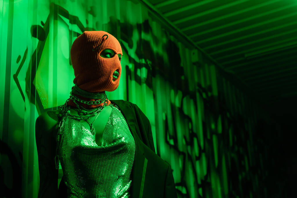 anonim szexi nő narancs símaszkban és ezüst felsőben, aki a fal felé néz graffitivel zöld fényben. - Fotó, kép