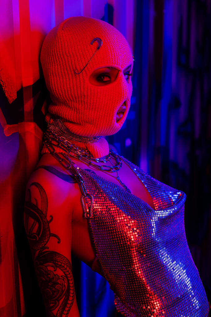 сексуальная женщина с татуировками в вязаной балаклаве и серебряном топе, смотрящая на камеру рядом с граффити на синем фоне при красном освещении - Фото, изображение