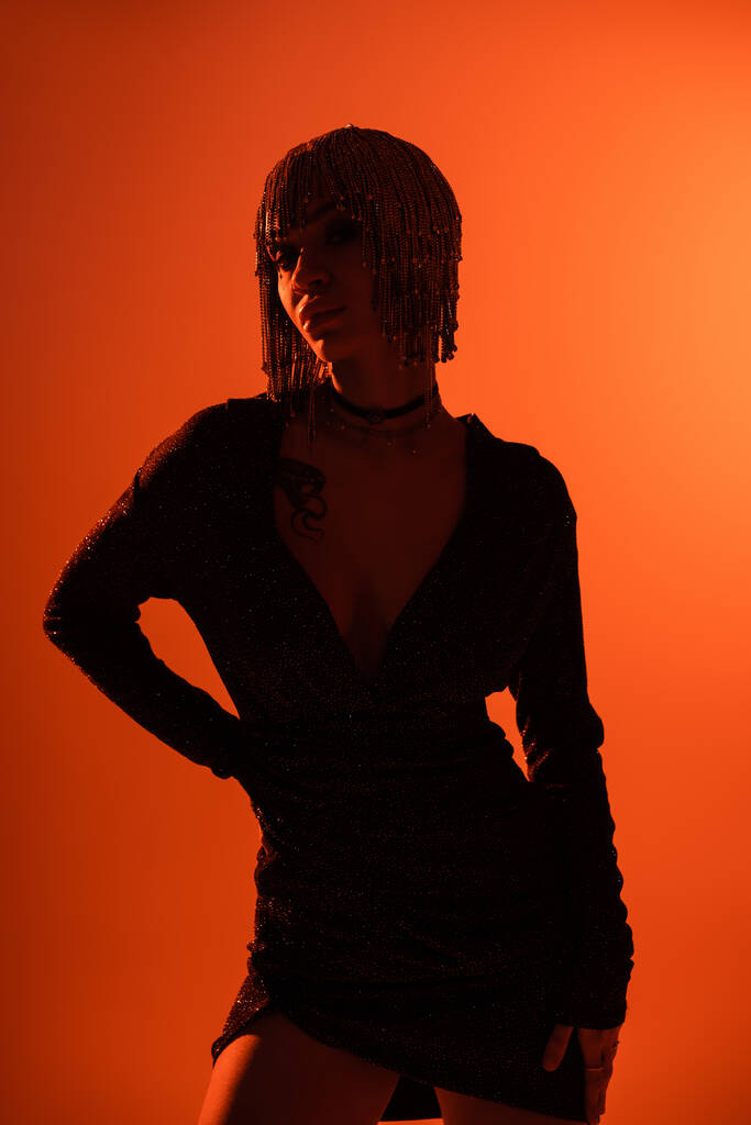 mujer tatuada en vestido negro y ropa de cabeza metálica posando con la mano en el fondo naranja cintura - Foto, imagen