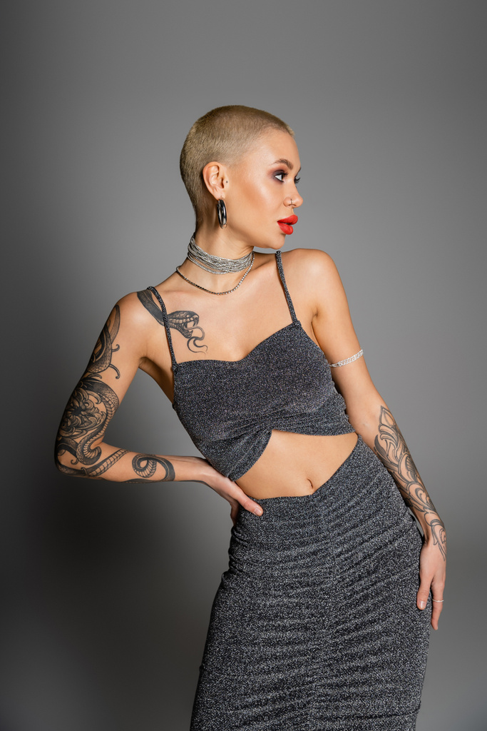 seductive woman in lurex crop top and necklaces looking away while posing with hand on hip on grey background - Photo, Image