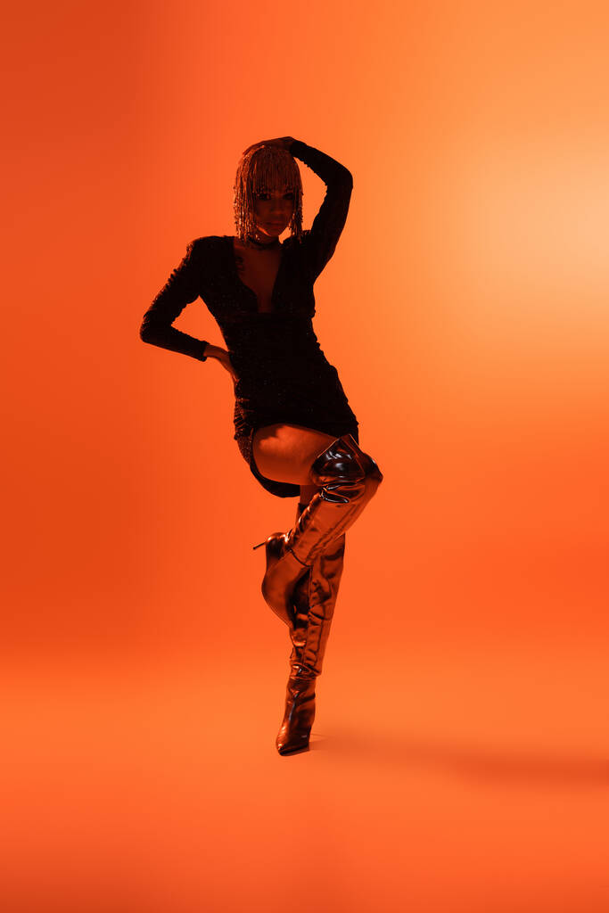 volledige lengte van sexy model in modieuze laarzen en zwarte jurk poseren op een been op oranje achtergrond - Foto, afbeelding
