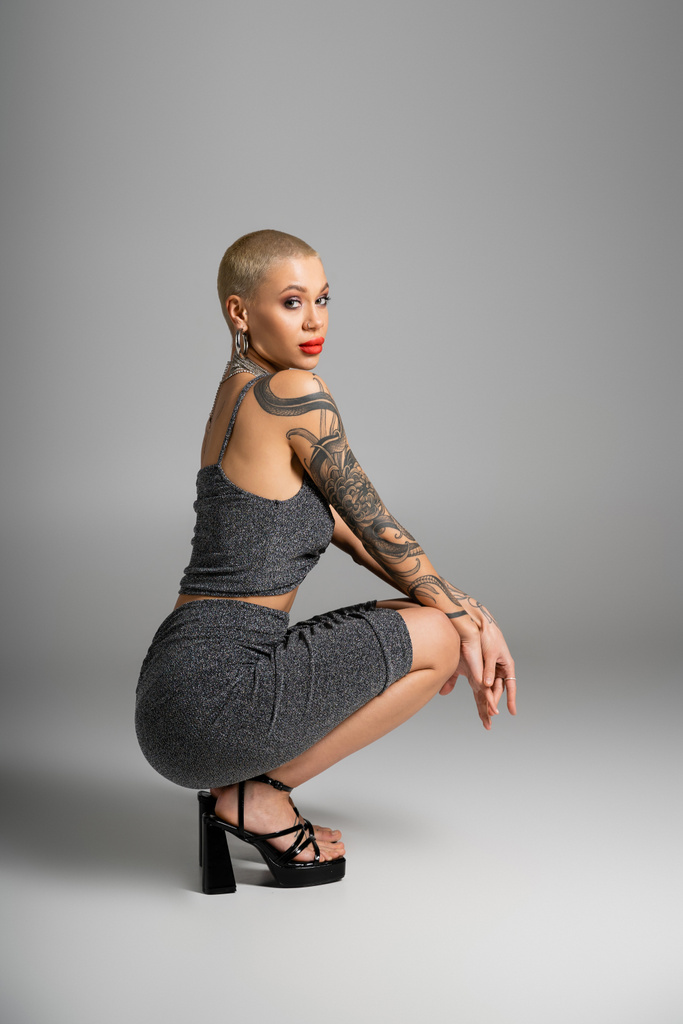 full length of sexy tattooed woman in lurex skirt and heeled sandals sitting on haunches on grey background - Photo, Image