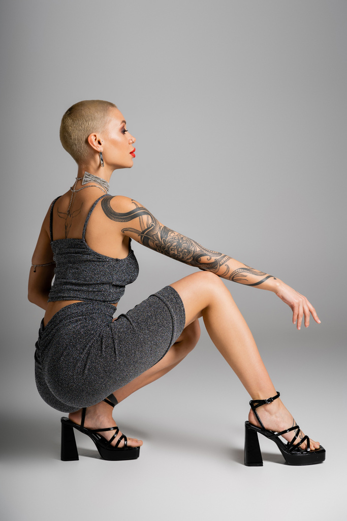 side view of tattooed glamour woman in sexy outfit sitting on haunches on grey background - Photo, Image