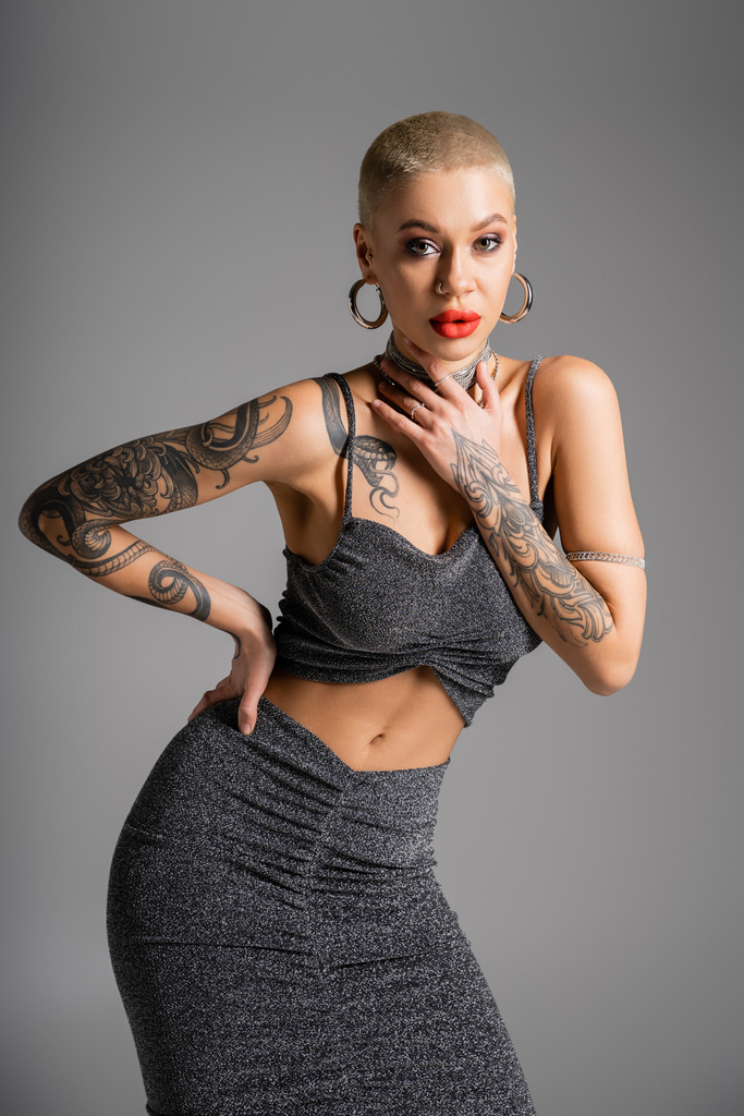 seductive tattooed woman in lurex skirt and crop top touching necklaces while posing with hand on hip isolated on grey - Photo, Image