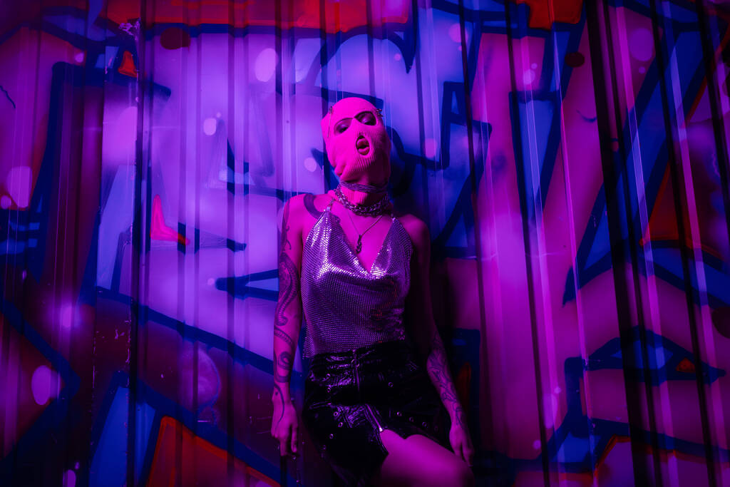 sexy woman in balaclava and silver top with black skirt posing near colorful graffiti in purple light - Photo, Image