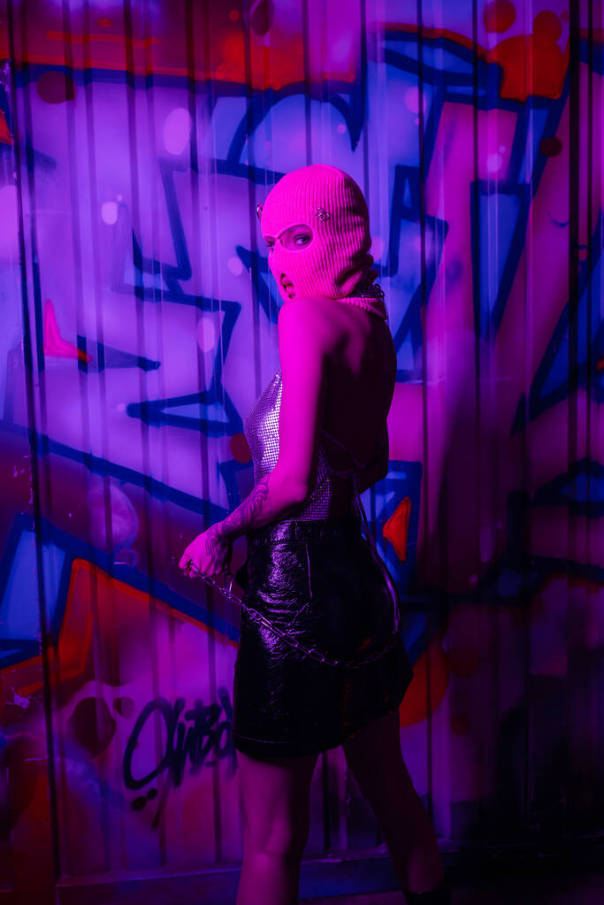 seductora mujer en pasamontañas rosa posando con cadena plateada y mirando a cámara cerca de pared con graffiti en iluminación púrpura - Foto, Imagen