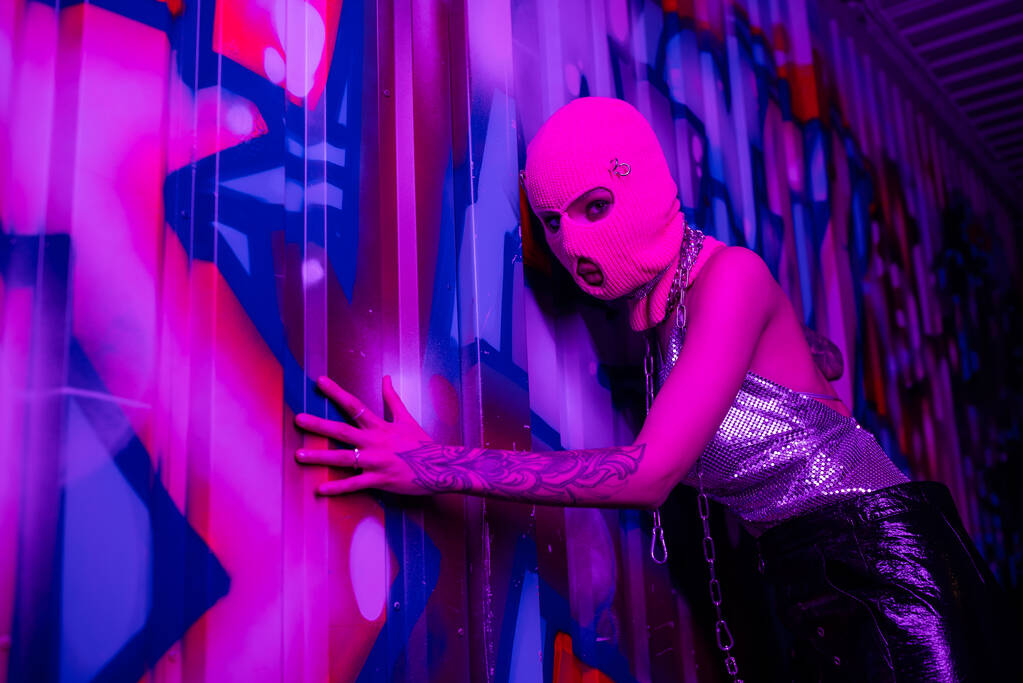 ανώνυμη γυναίκα με τατουάζ σε balaclava συγκινητικό τοίχο με πολύχρωμα γκράφιτι και κοιτάζοντας κάμερα σε μωβ νέον φως - Φωτογραφία, εικόνα