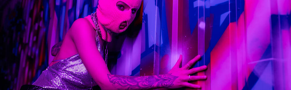 сексуальная женщина в розовой балаклаве смотрит в камеру, прикасаясь к красочным граффити в фиолетовом свете, баннер - Фото, изображение