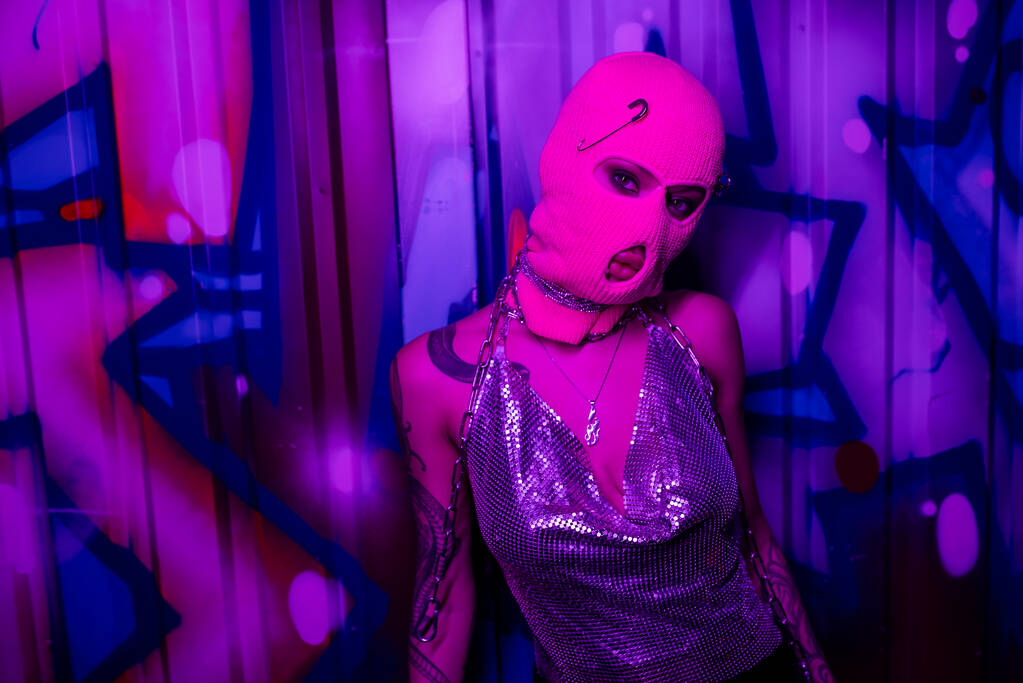 verführerische tätowierte Frau in metallisch glänzendem Oberteil und pinker Sturmhaube, die in Wandnähe steht, mit Graffiti in lila Neonlicht - Foto, Bild